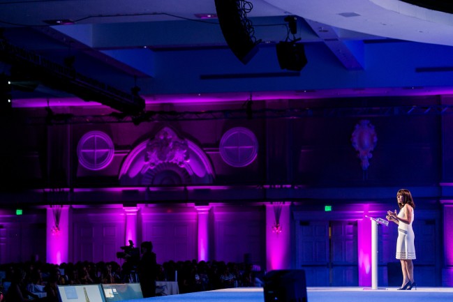 woman in white dress on stage addresses a corporate event audience